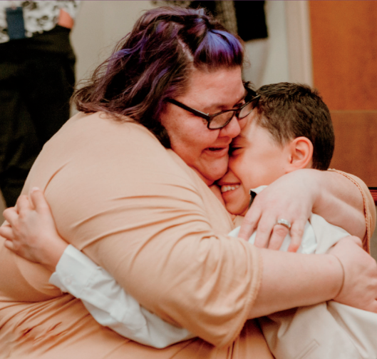 A woman with purple hair and glasses hugs a boy tightly, both smiling with eyes closed in a warm embrace. The woman is wearing a beige top, and the boy is in a light-colored outfit. They lean their heads together, expressing love and affection.