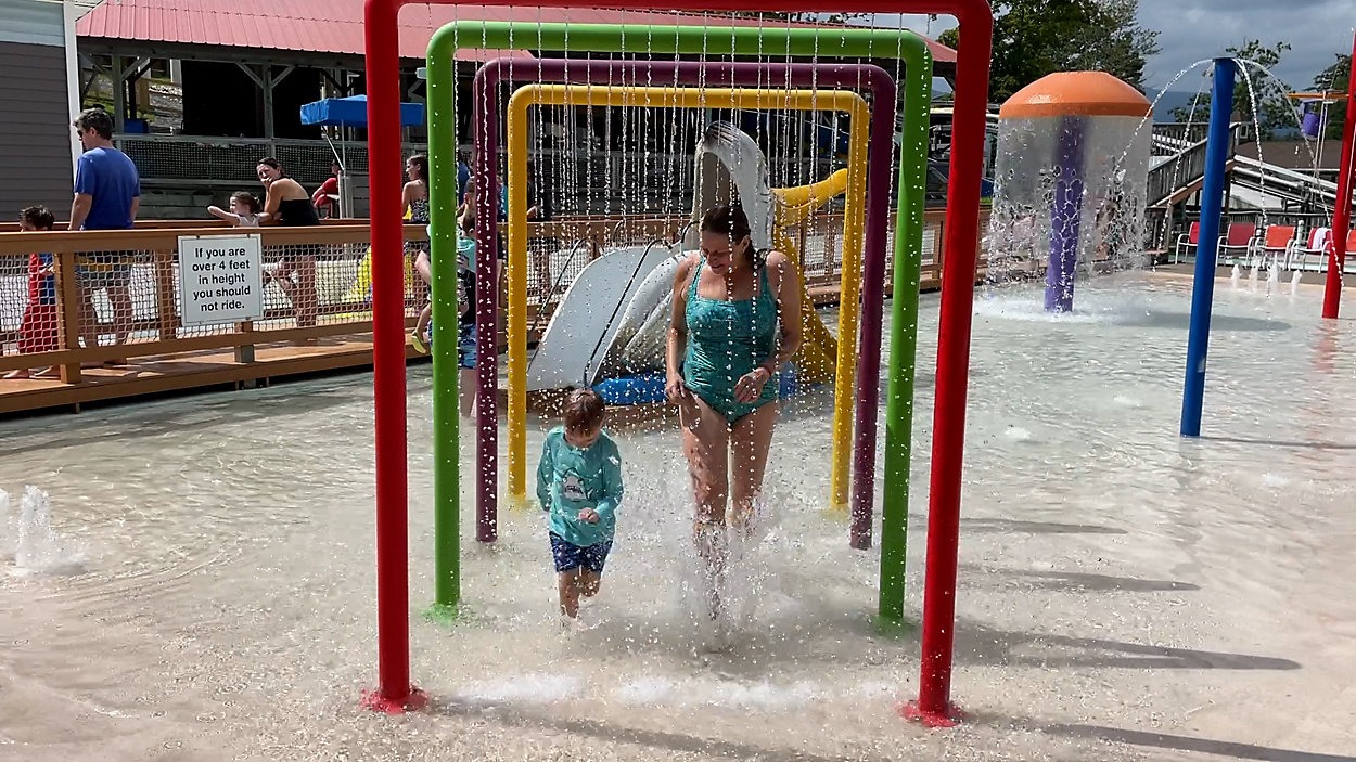 A woman and a child are walking through a series of water arches at a splash pad. They are both in swimsuits and appear to be smiling. In the background, there are water slides, a mushroom-shaped water feature, and a few people enjoying the water area.