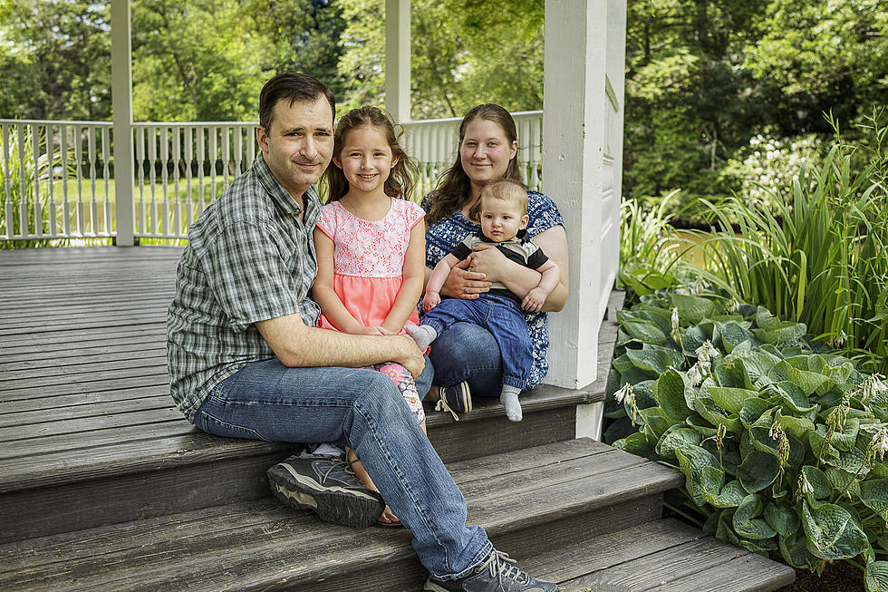 A family of four sits together on the steps of a gazebo. The father and daughter sit to the left, and the mother holds a baby boy on the right. They are surrounded by greenery, and the gazebo is situated in a lush, outdoor setting.