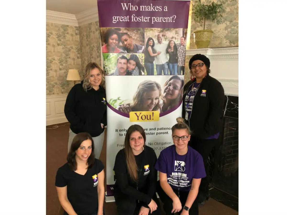 A group of five people poses in front of a banner with the text "Who makes a great foster parent? You!" They are indoors, and the banner features images of smiling families. Four individuals are standing, and one is kneeling in front. They all wear matching outfits.