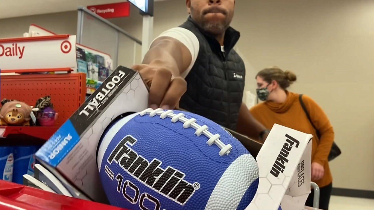 A person reaches towards a blue Franklin youth football displayed on a store shelf. Another person, wearing a face mask, walks past in the background. Various store items and signage are visible nearby.