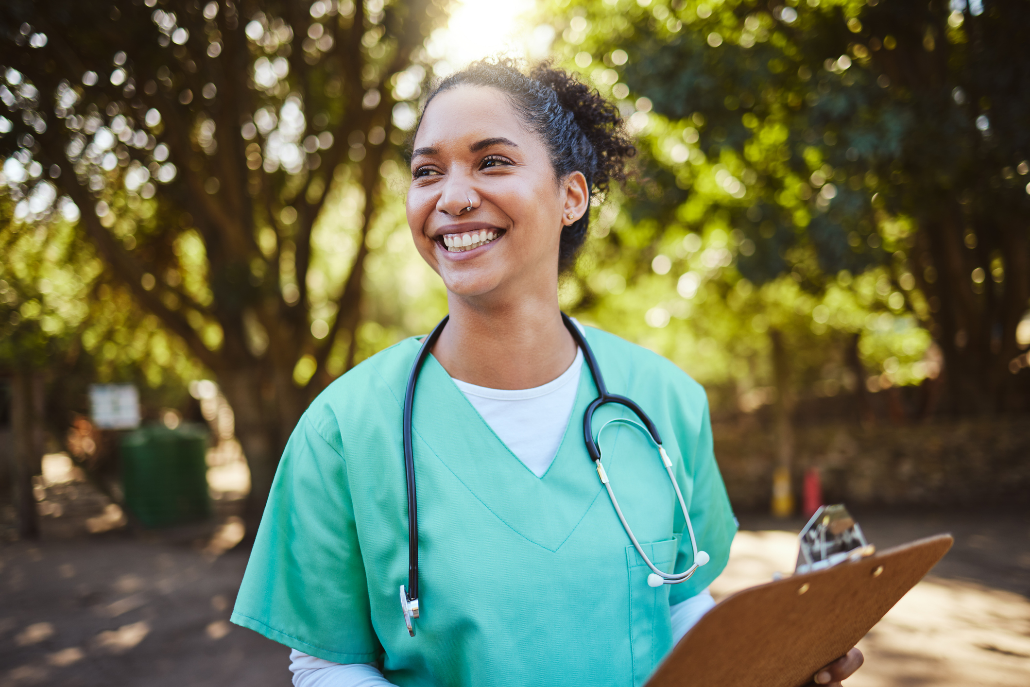 A smiling woman wearing green medical scrubs and a stethoscope around her neck holds a clipboard. She has curly hair pulled back and stands outdoors with trees in the background, illuminated by sunlight.