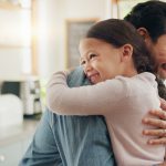 A father embraces his young daughter in a warm hug in a bright, modern kitchen. Both are smiling joyfully. The kitchen features shelves with plants, a microwave, and sunlight streaming through the windows, adding a warmth to the scene.