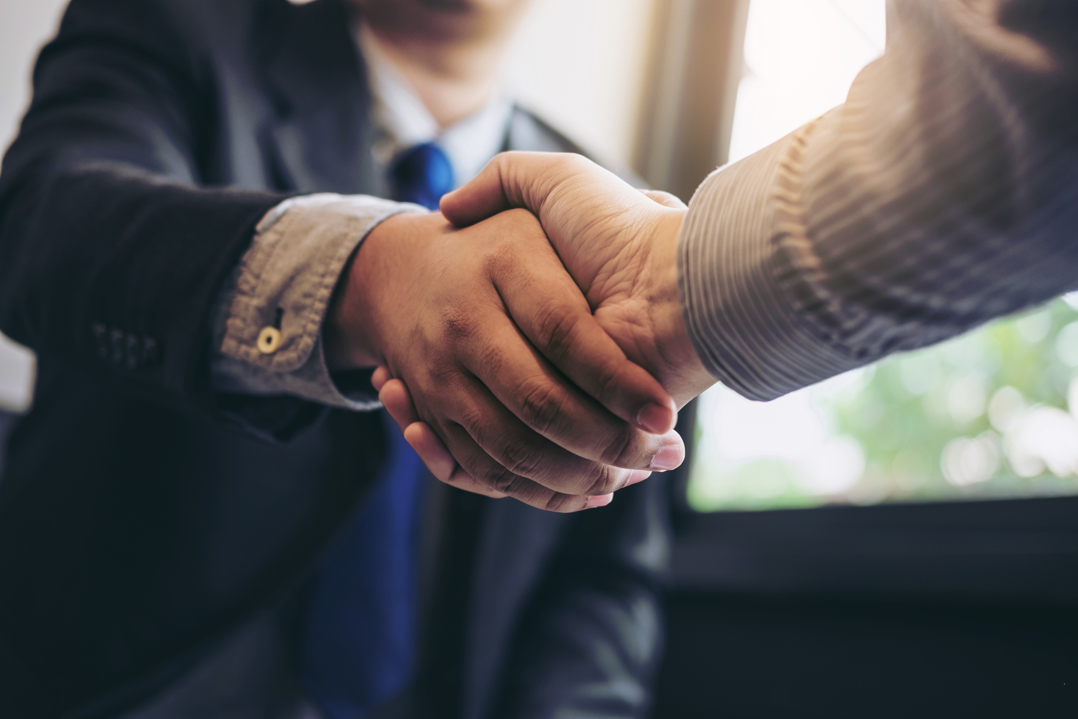 Two people in business attire shaking hands, symbolizing an agreement or partnership. The background is slightly blurred, with natural light coming through a window, giving a warm tone to the scene.