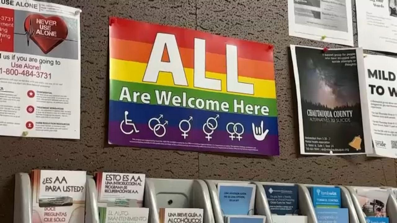 A wall-mounted poster with a rainbow background reads "ALL Are Welcome Here," featuring inclusive symbols for accessibility, gender, sexual orientation, and American Sign Language for "I love you." Below are various brochures and pamphlets in wall-mounted holders.