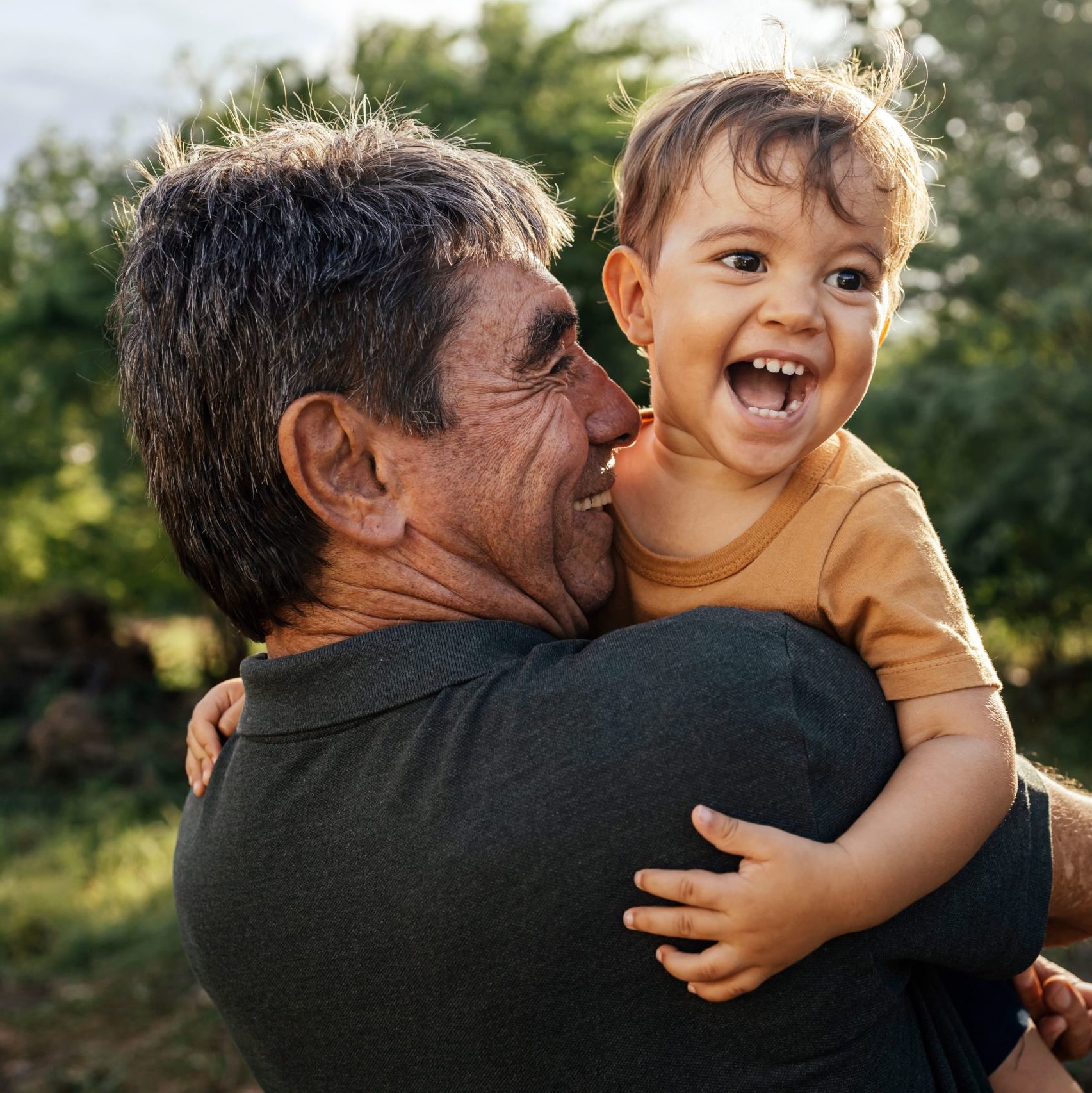 A grandfather holding his joyful grandson outdoors. The man has graying hair and wears a dark shirt, while the child has light brown hair and wears a mustard-colored shirt. They both smile widely, standing in a natural setting with greenery in the background.