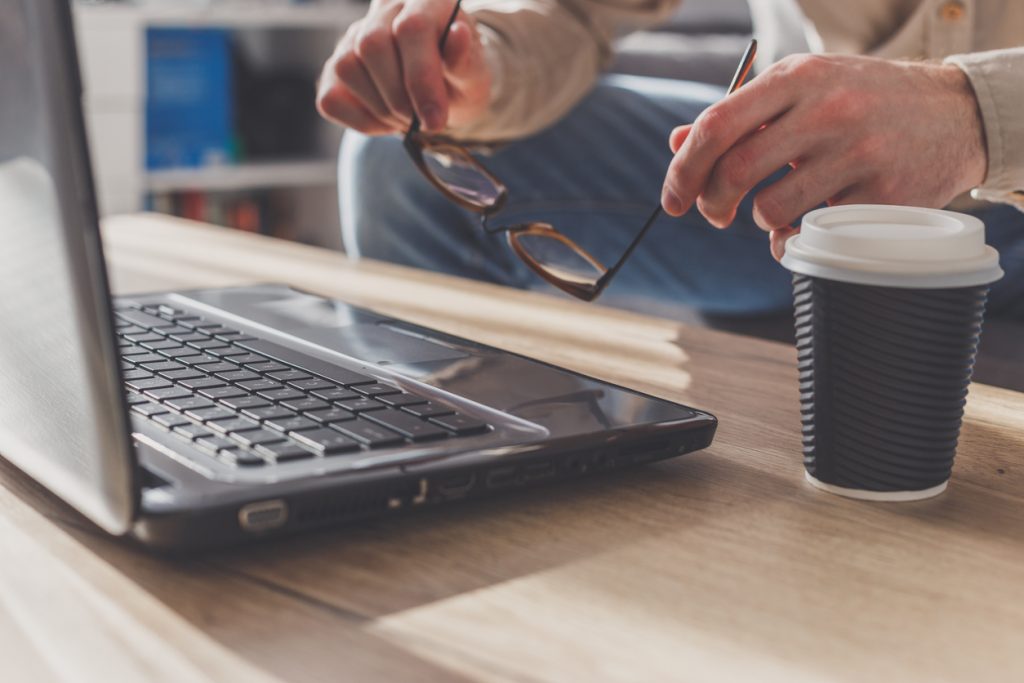 Man putting up eyeglasses on laptop to make a short break, tired after long work or getting nervous. Coffee cup on table near laptop.