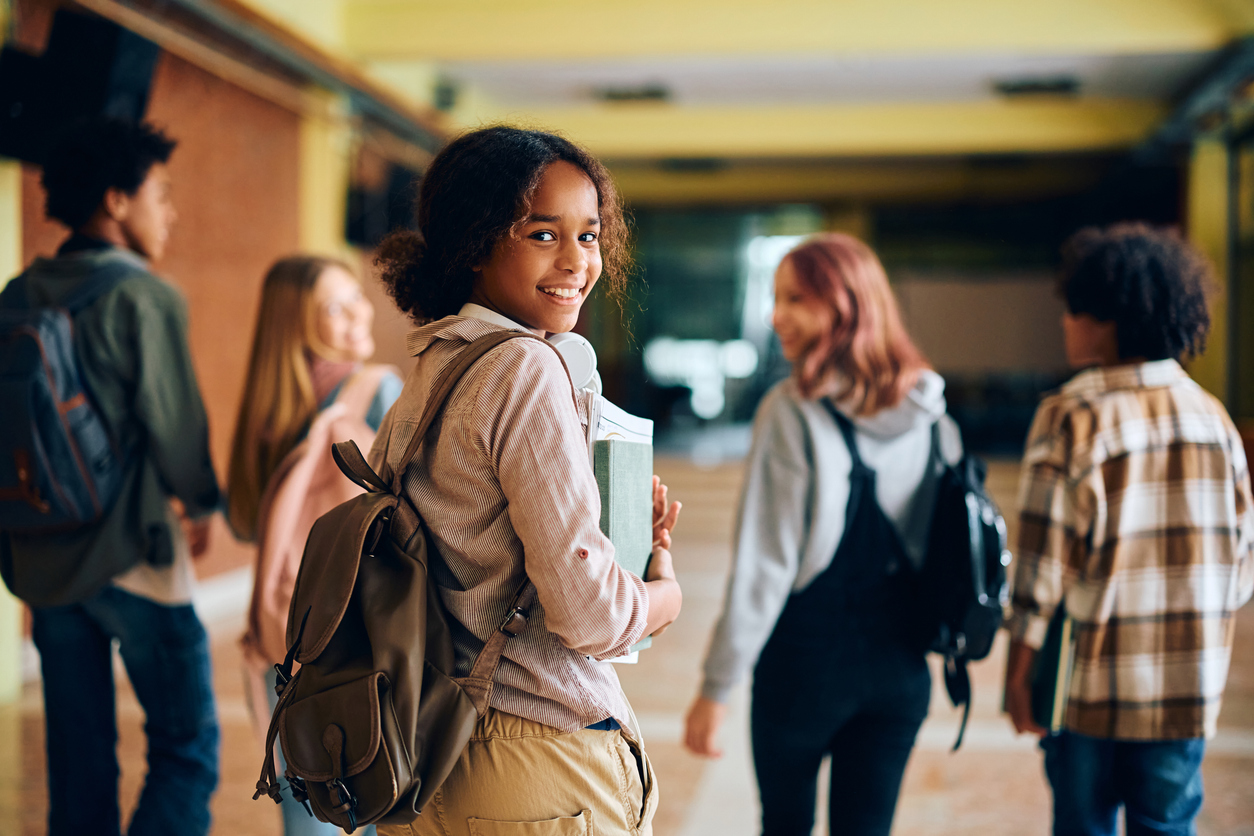 A smiling student with curly hair, wearing a backpack and holding notebooks, looks back while walking down a school hallway. Four other students in casual clothes and backpacks walk ahead, engaged in conversation. The hallway is brightly lit.