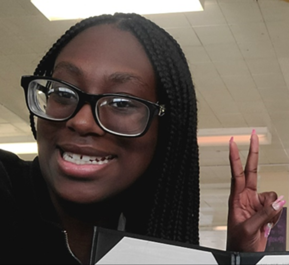 A person with braided hair and glasses smiles while holding up a book. They are making a peace sign with their other hand. The background appears to be an indoor setting with fluorescent lighting and ceiling tiles.