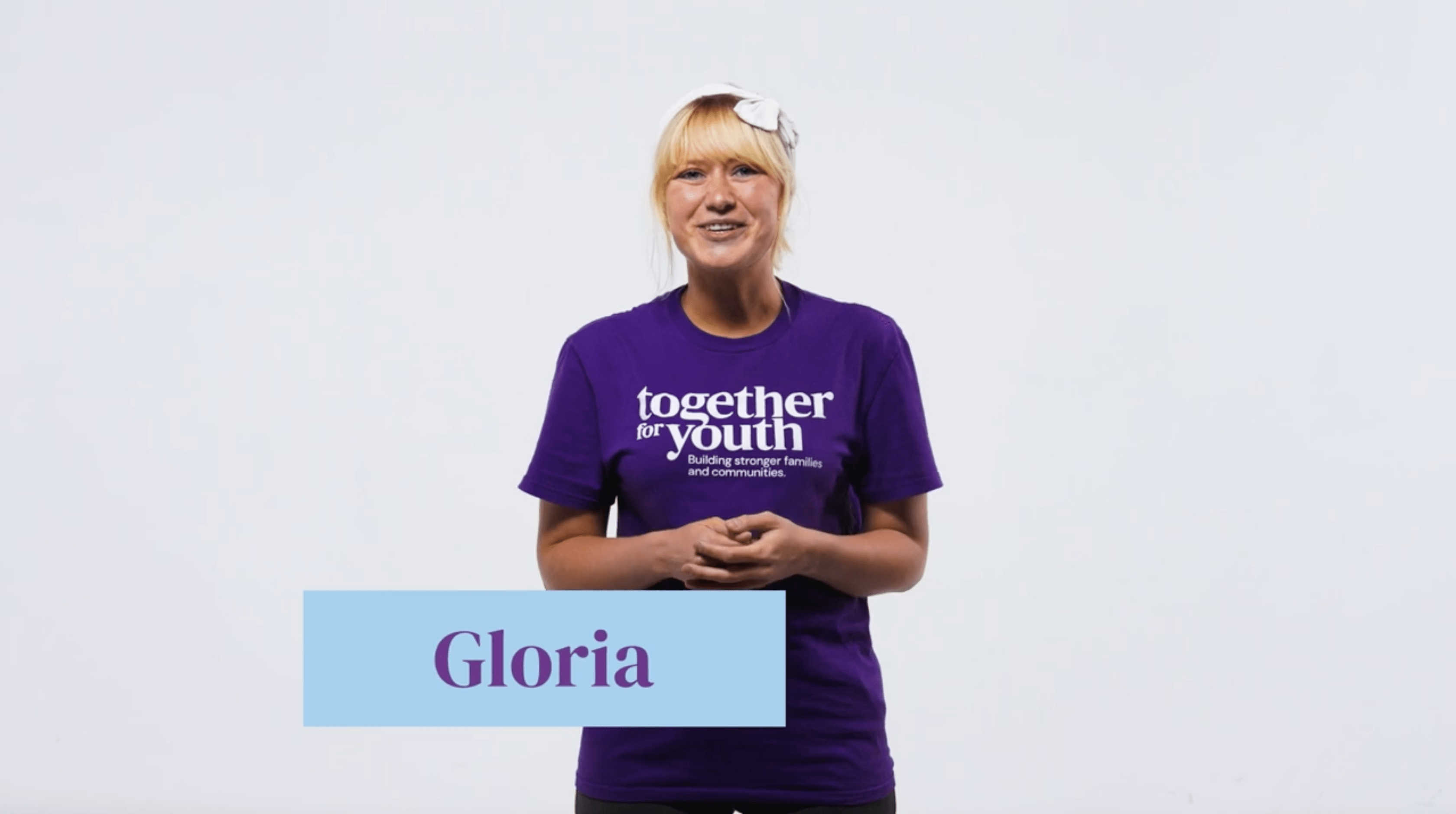 Gloria with blonde hair stands against a plain background, smiling and wearing a purple shirt that reads, 