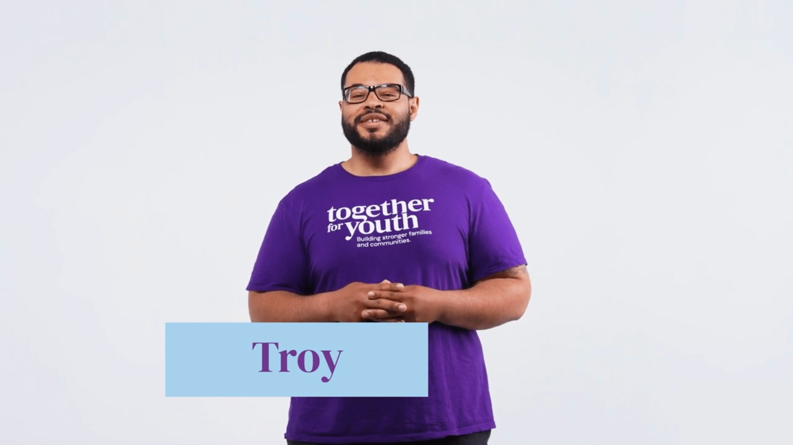 Troy is pictured with a beard and glasses, and stands smiling against a light background. He is wearing a purple t-shirt with the text 