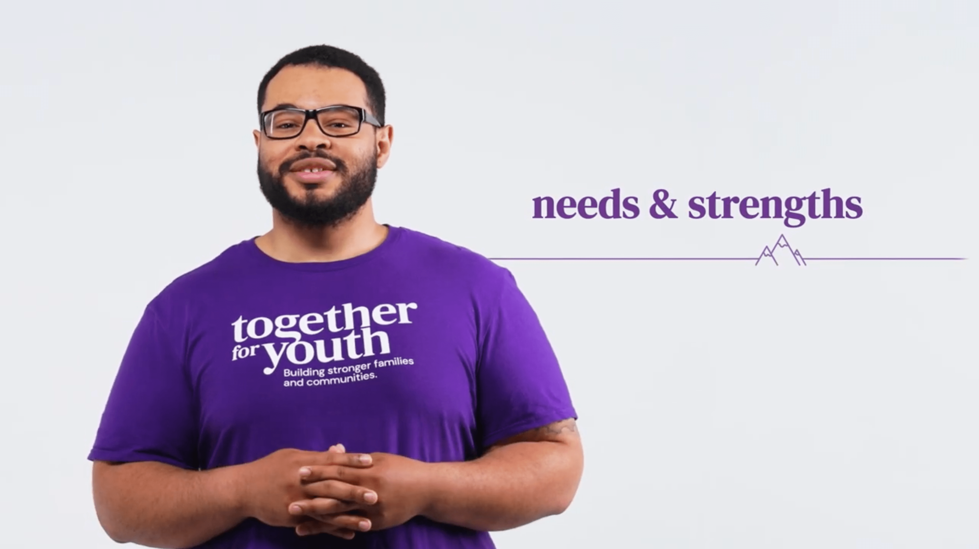 Troy is pictured with glasses and a beard. He stands against a white background. He is wearing a purple t-shirt that reads 