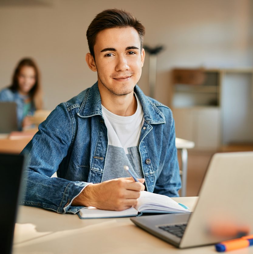 A young man with short brown hair, wearing a denim jacket, sits at a desk with a laptop and notebook. He is smiling and holding a pen. In the background, two people are working on laptops in a well-lit room.