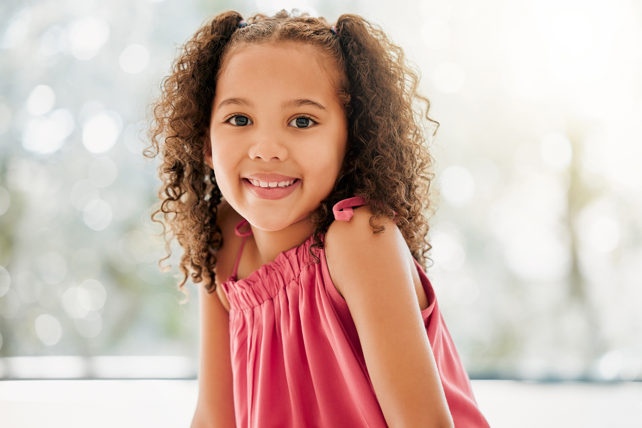 A young girl with curly hair smiles warmly at the camera. She is wearing a pink sleeveless top. The background is softly blurred, giving a bokeh effect with gentle light shining through, creating a bright and cheerful atmosphere.