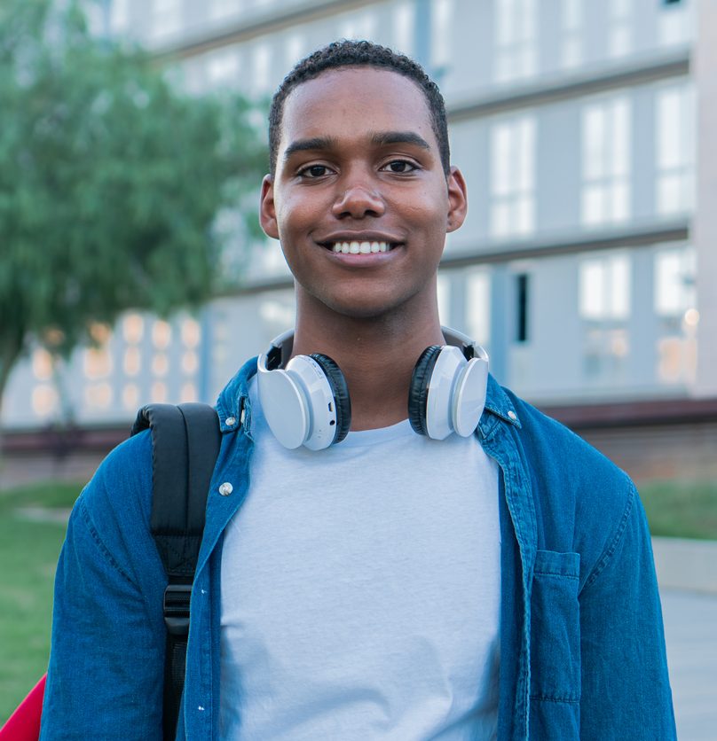 A young man stands outside a modern building, smiling at the camera. He wears a blue denim shirt over a white t-shirt, headphones around his neck, and carries a backpack. A blurry tree and grassy area are in the background.