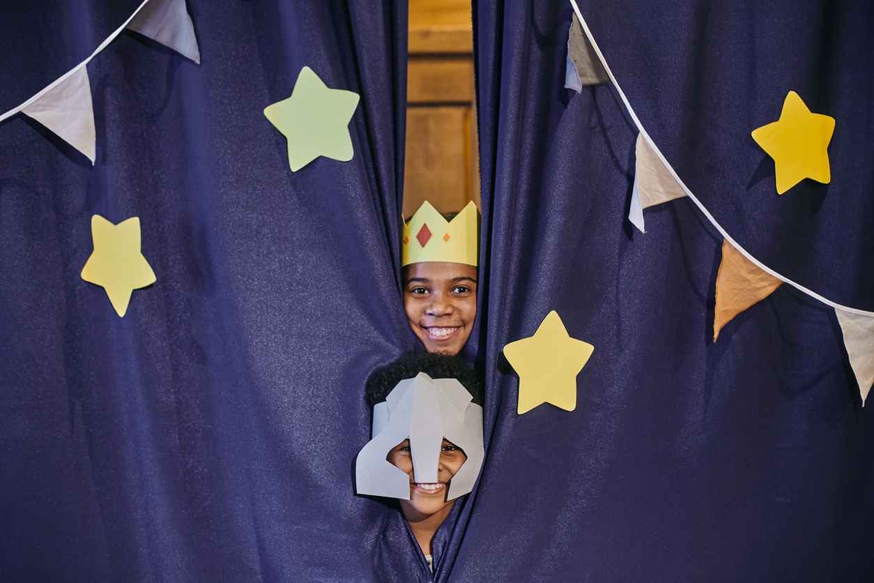 Two children peek through a dark blue curtain adorned with star and triangle cutouts. The child on top wears a yellow crown, while the child below sports a gray helmet. Both are smiling brightly, bringing a playful and joyful atmosphere to the scene.