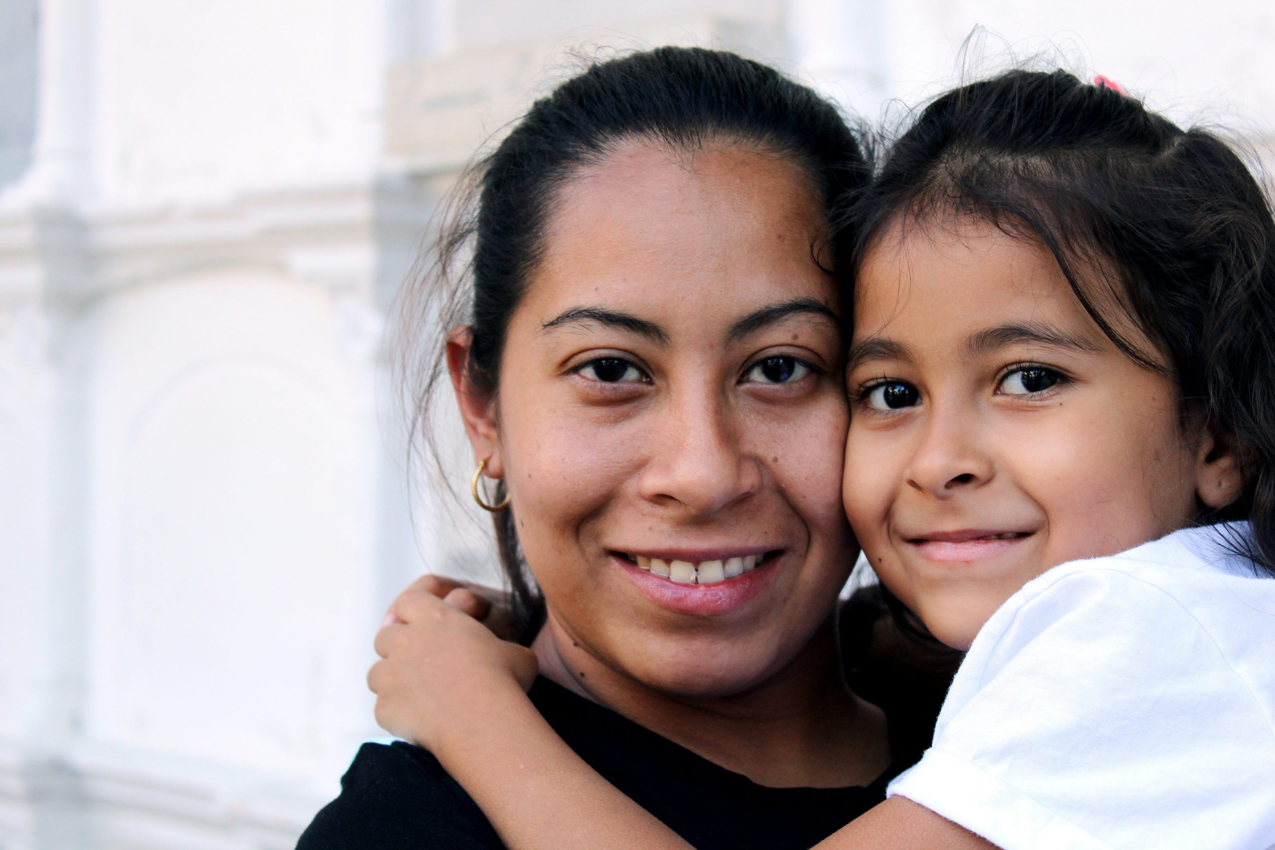 A close-up of a woman and a young girl smiling at the camera. The girl is being held by the woman and has her arms around the woman's neck. Both appear happy and are standing against a light-colored, blurry background.