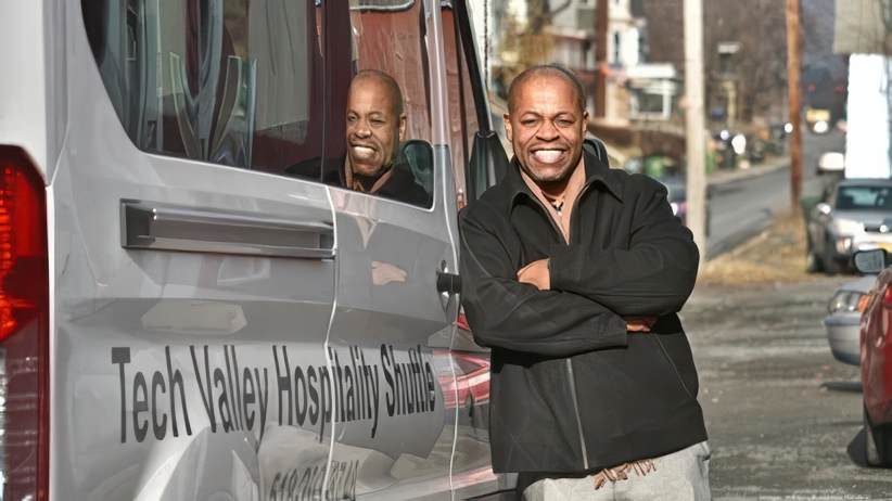 A man stands smiling with arms crossed next to a van labeled "Tech Valley Hospitality Shuttle." His reflection is visible in the van window. The scene is outdoors, with a street and buildings in the background.