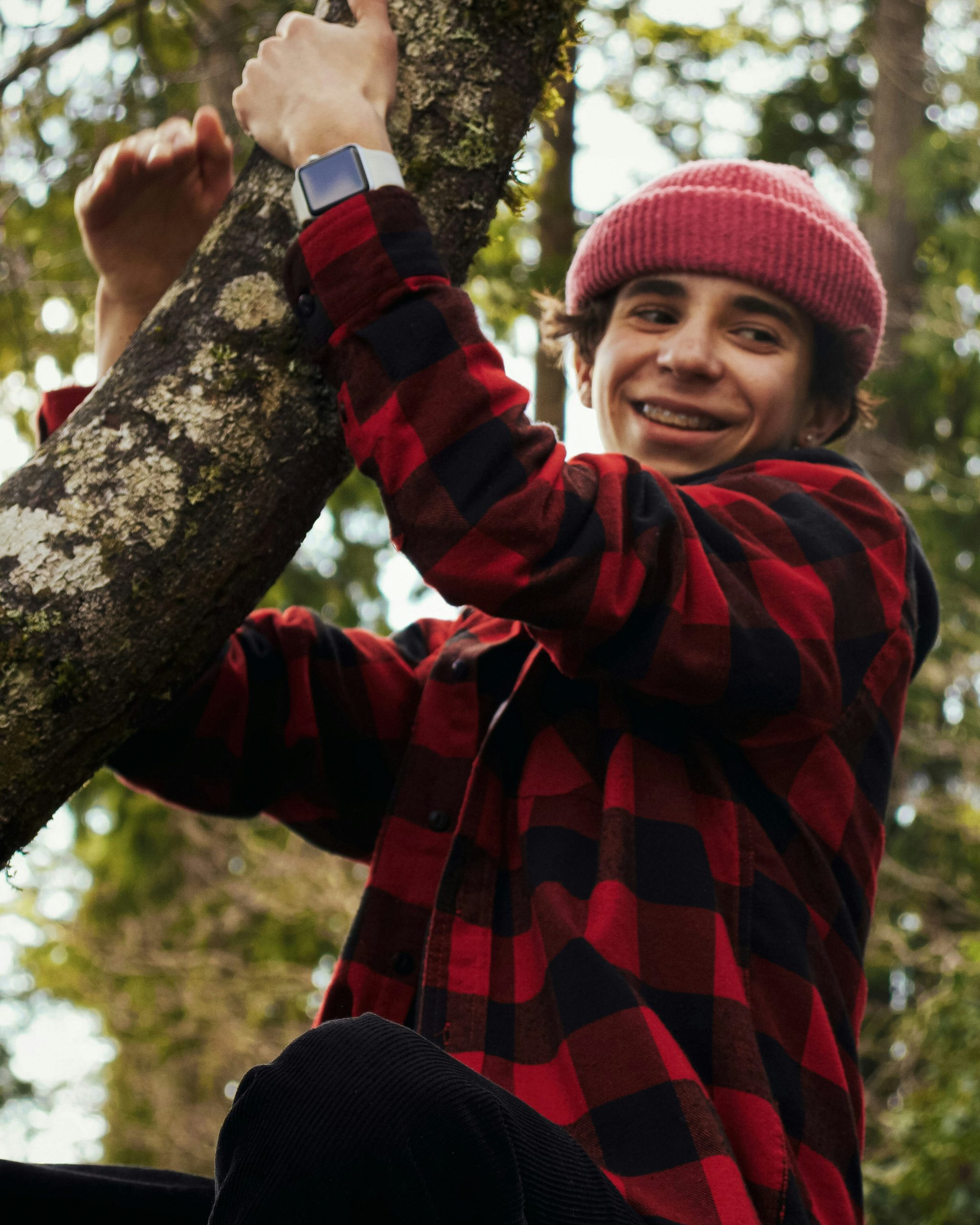 A person wearing a red and black checkered shirt and a pink beanie smiles while climbing a tree. They wear a watch on their wrist, and the background is filled with greenery.