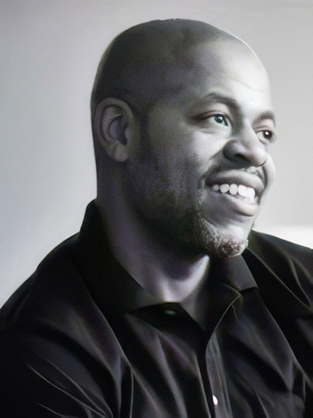 A smiling man with a beard and a shaved head is looking off to the side. He is wearing a dark collared shirt. The background is softly blurred, giving a professional portrait feel.