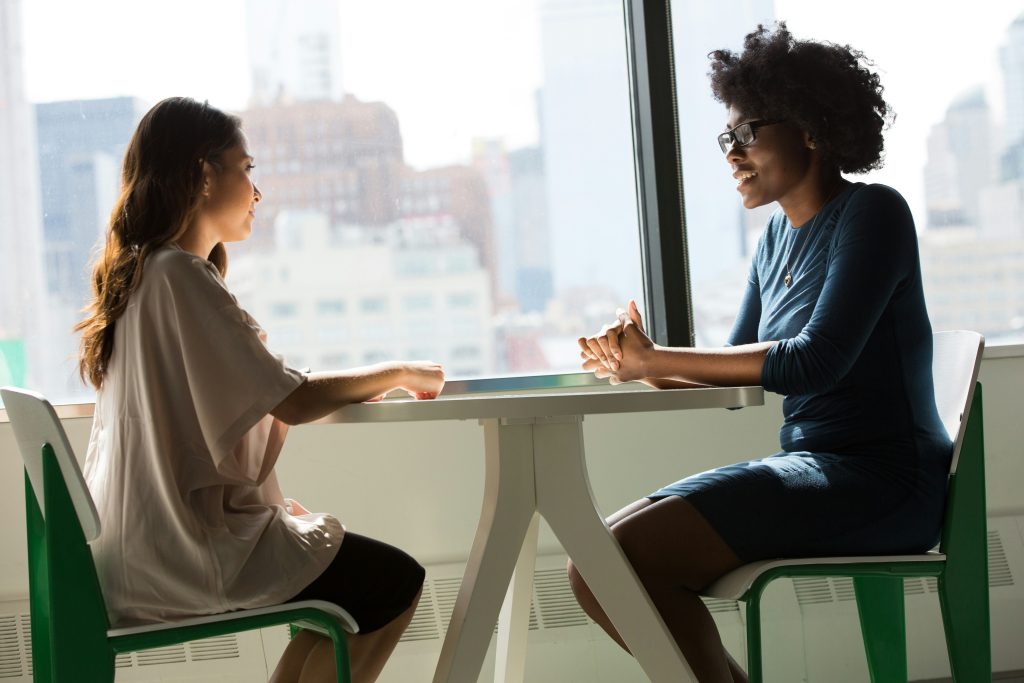 Two women sit across from each other at a table, engaged in conversation. One woman wears a light blouse, the other a dark dress. They are near a large window with an urban cityscape in the background.