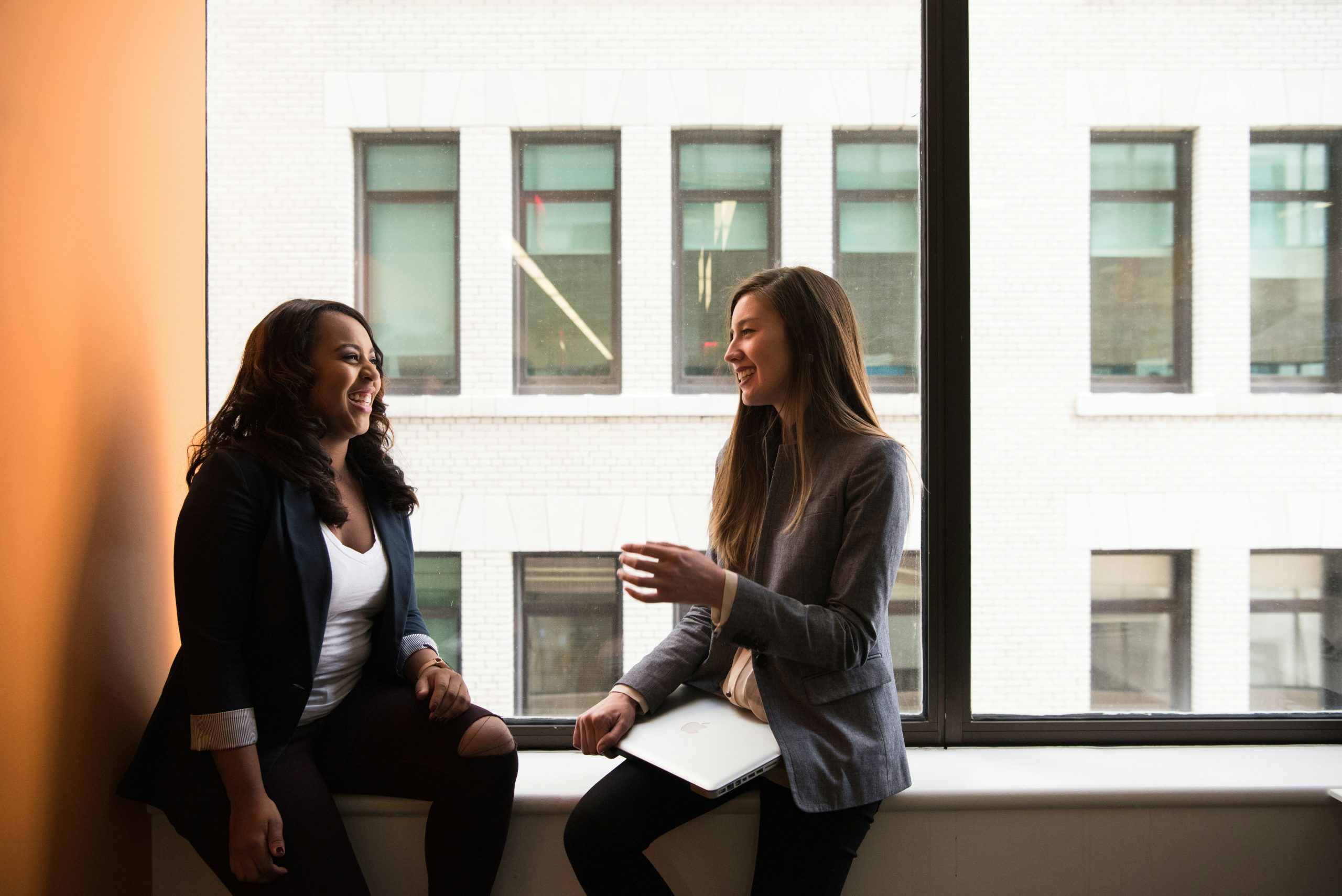 Two women are sitting on a windowsill in an office, engaged in conversation. Both are dressed in business attire. One holds a laptop, and they appear to be smiling, creating a friendly and professional atmosphere.