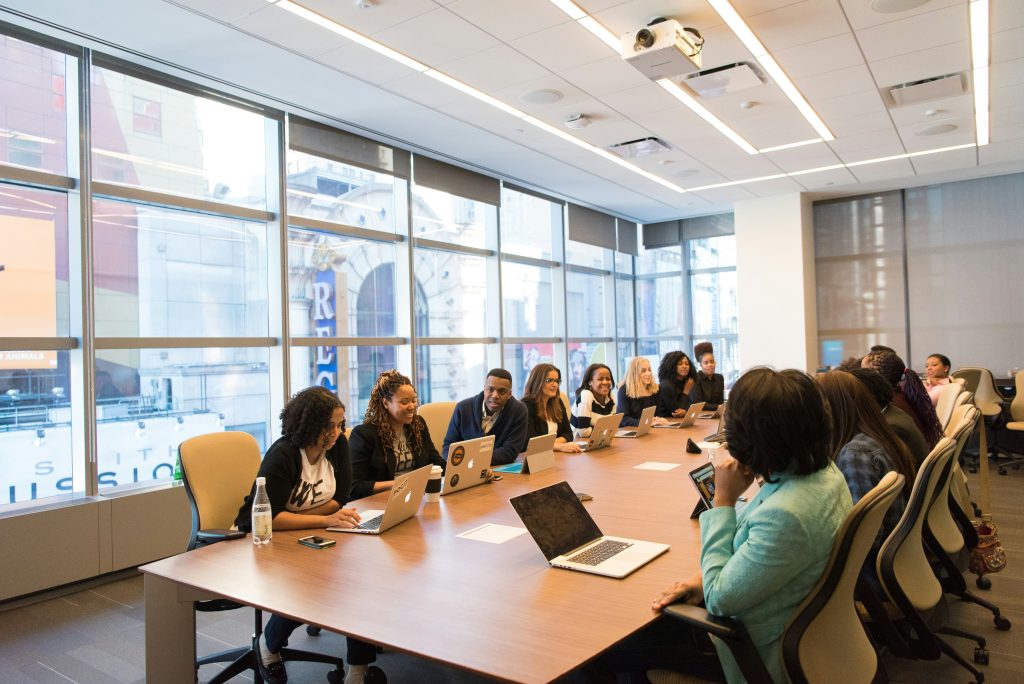 A diverse group of people is sitting around a large conference table in a modern office setting. They are engaged in a meeting, using laptops, with large windows in the background showing an urban view.