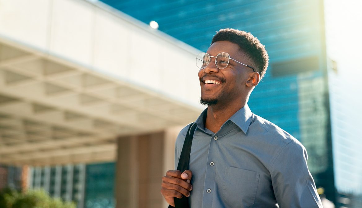 A man wearing glasses and a blue shirt smiles while walking outside in the sunlight. He carries a bag over his shoulder. A modern building and clear sky are in the background.