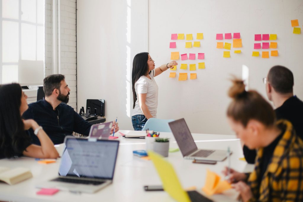 A woman stands at a whiteboard covered with colorful sticky notes, pointing to one. She is in a meeting room with four colleagues seated around a table, each with laptops and notes, engaging in a discussion.