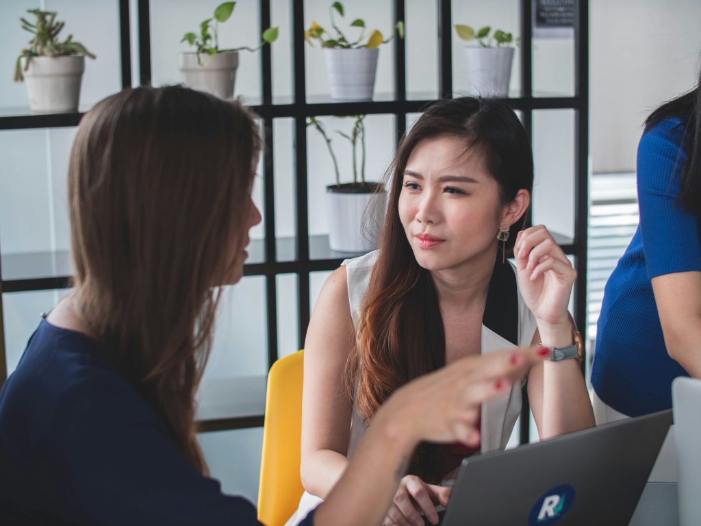 Two women are sitting at a table, engaged in conversation. One is gesturing while the other listens attentively with a laptop open in front of her. In the background, there is a shelf with potted plants.