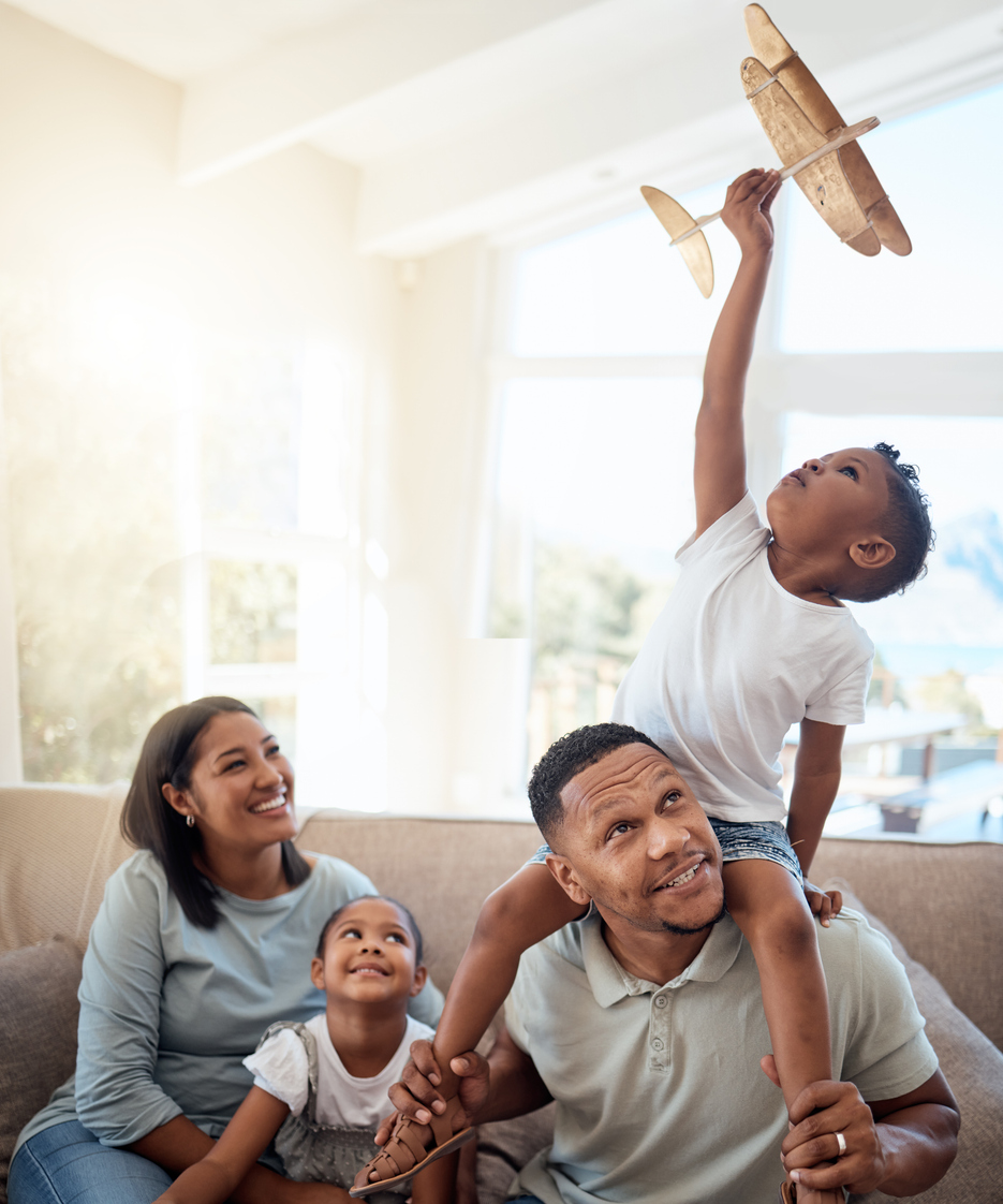 A family of four enjoys time together indoors. The young boy sits on the father's shoulders, holding up a toy airplane. The mother and daughter sit on the couch, smiling and watching, with sunlight streaming through the window.