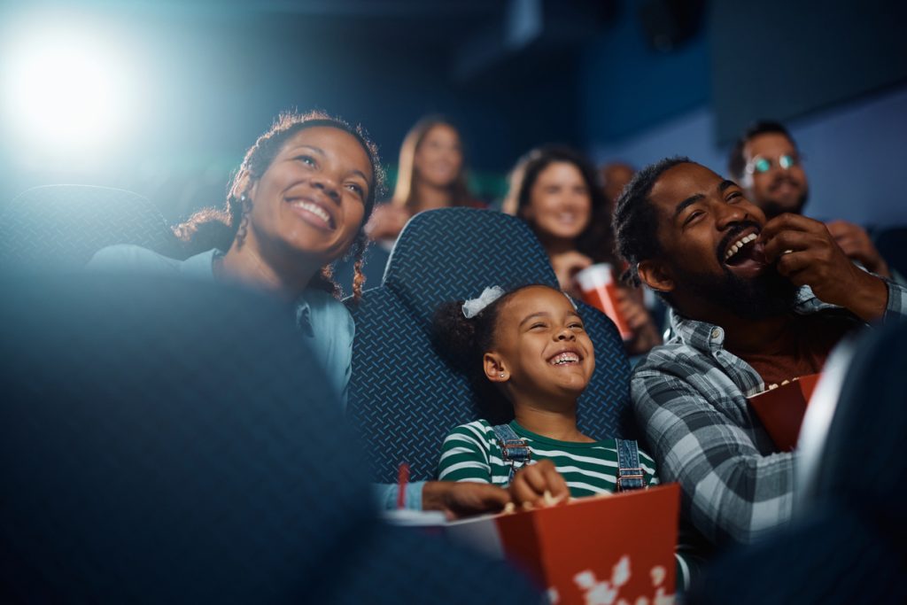 A diverse group of people in a movie theater enjoy a film. They are smiling and laughing while eating popcorn. The room is dimly lit, with focus on a family sitting together in cozy seats.