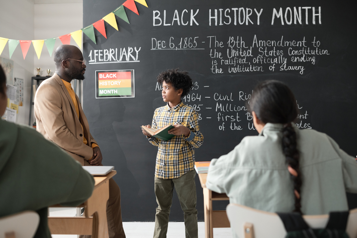 A teacher and student stand in a classroom decorated for Black History Month. The student holds a book and faces the teacher. A blackboard displays historical dates and events. Colorful flags hang above. Students sit at desks, viewing the interaction.