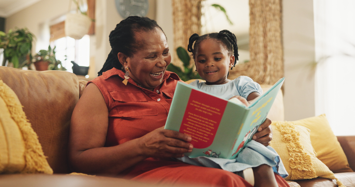 Grandmother and granddaughter sitting on a cozy couch, smiling while reading a colorful book together. The room has a warm ambiance with plants and soft pillows in the background.